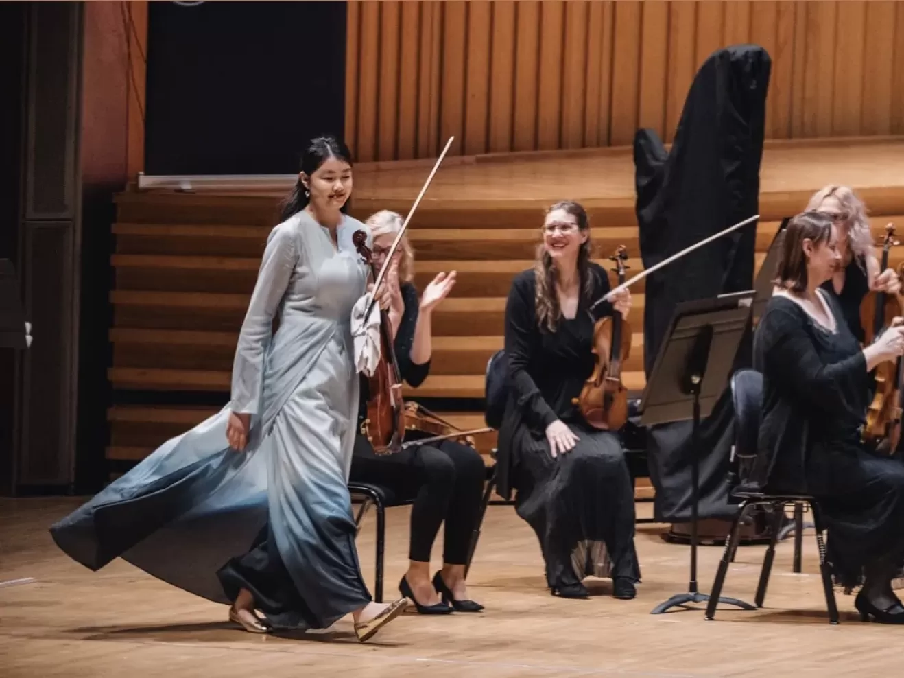 Xunyu Zhou läuft auf die Bühne mit ihrer Viola. Hinter ihr sitzen Orchestermusikerinnen, die teilweise lachen und klatschen.