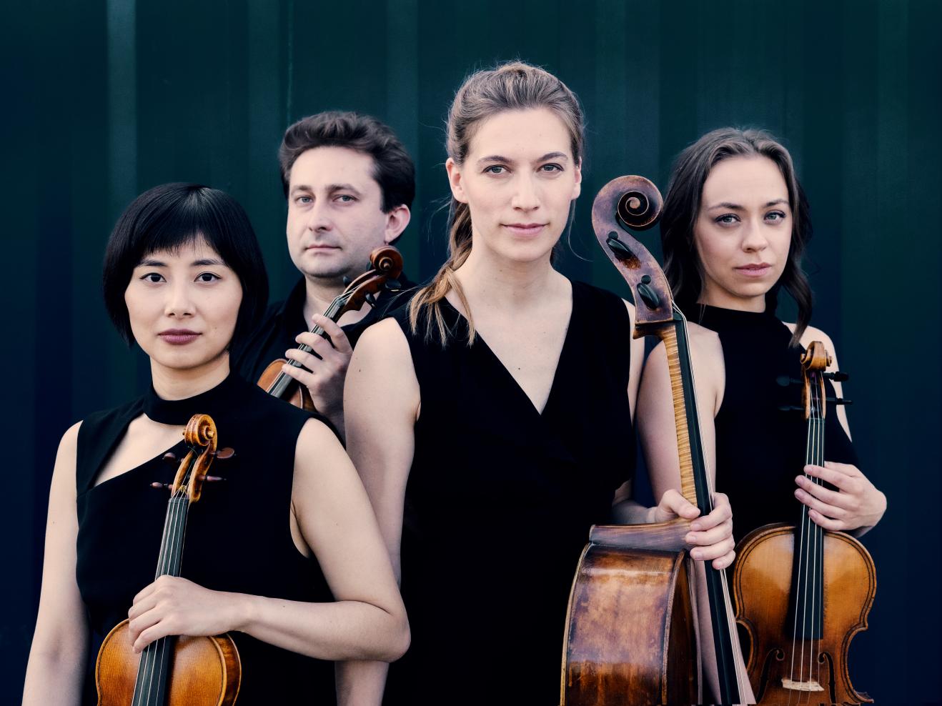 Portraitfoto des Ensembles: Die vier Musiker*innen tragen schwarze Kleidung und halten ihre Instrumente in den Händen. Sie stehen vor einem blauen Hintergrund und blicken eindringlich in die Kamera.