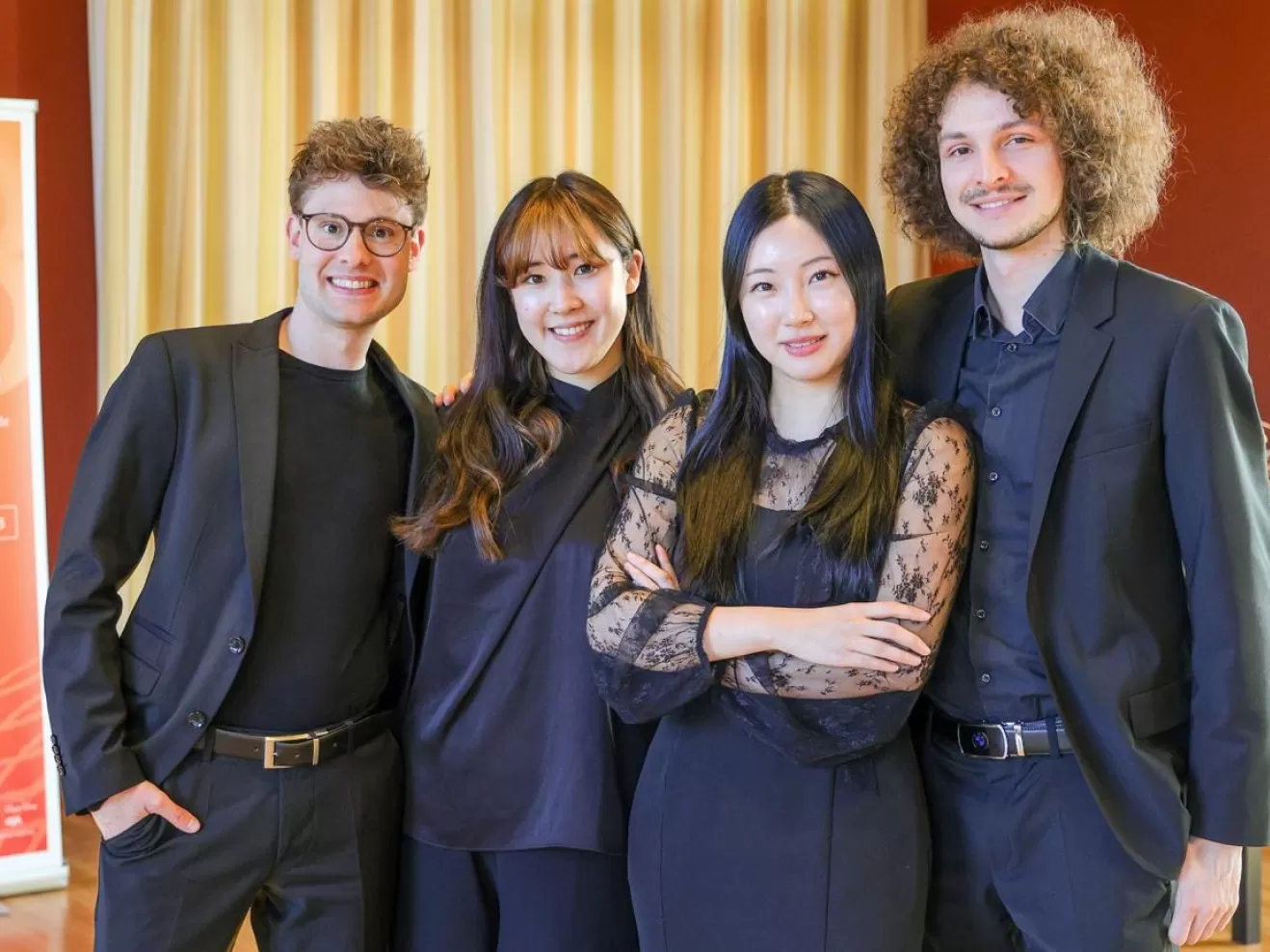 Die vier schwarz gekleideten Studierenden stehen vor einem Roll-up des Wettbewerbs und lächeln.