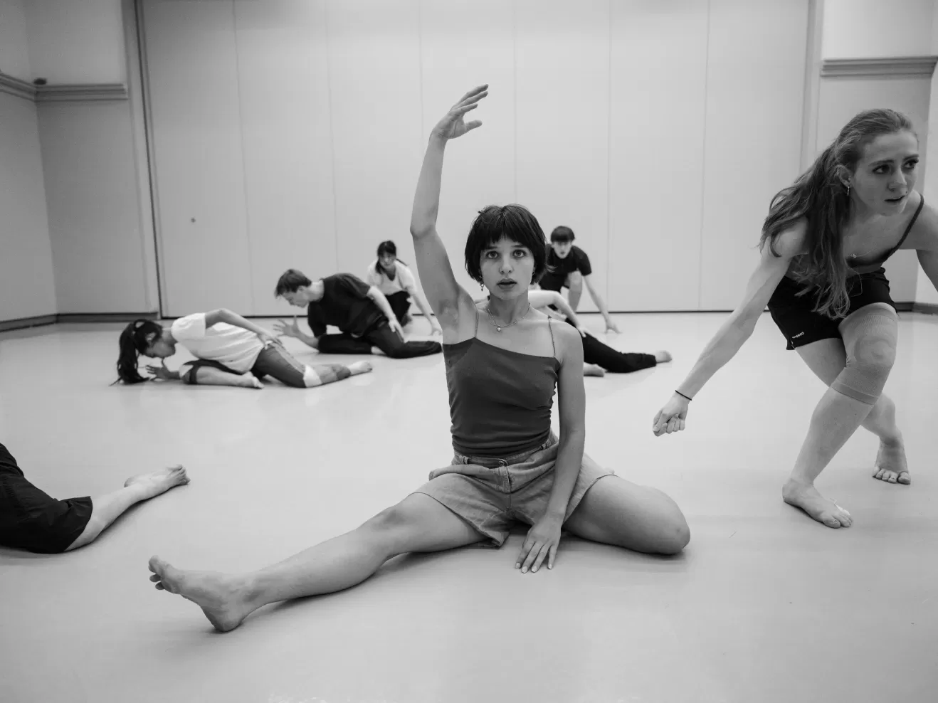 Schwarz-Weiß Bild einer Tanzprobe. Im Vordergrund eine Tänzerin, die sitzend direkt in die Kamera blickt und ihren Arm über den Kopf hebt.