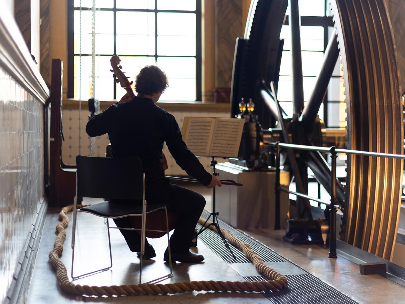 Ein Cellist spielt neben einem großen Zahnrad.