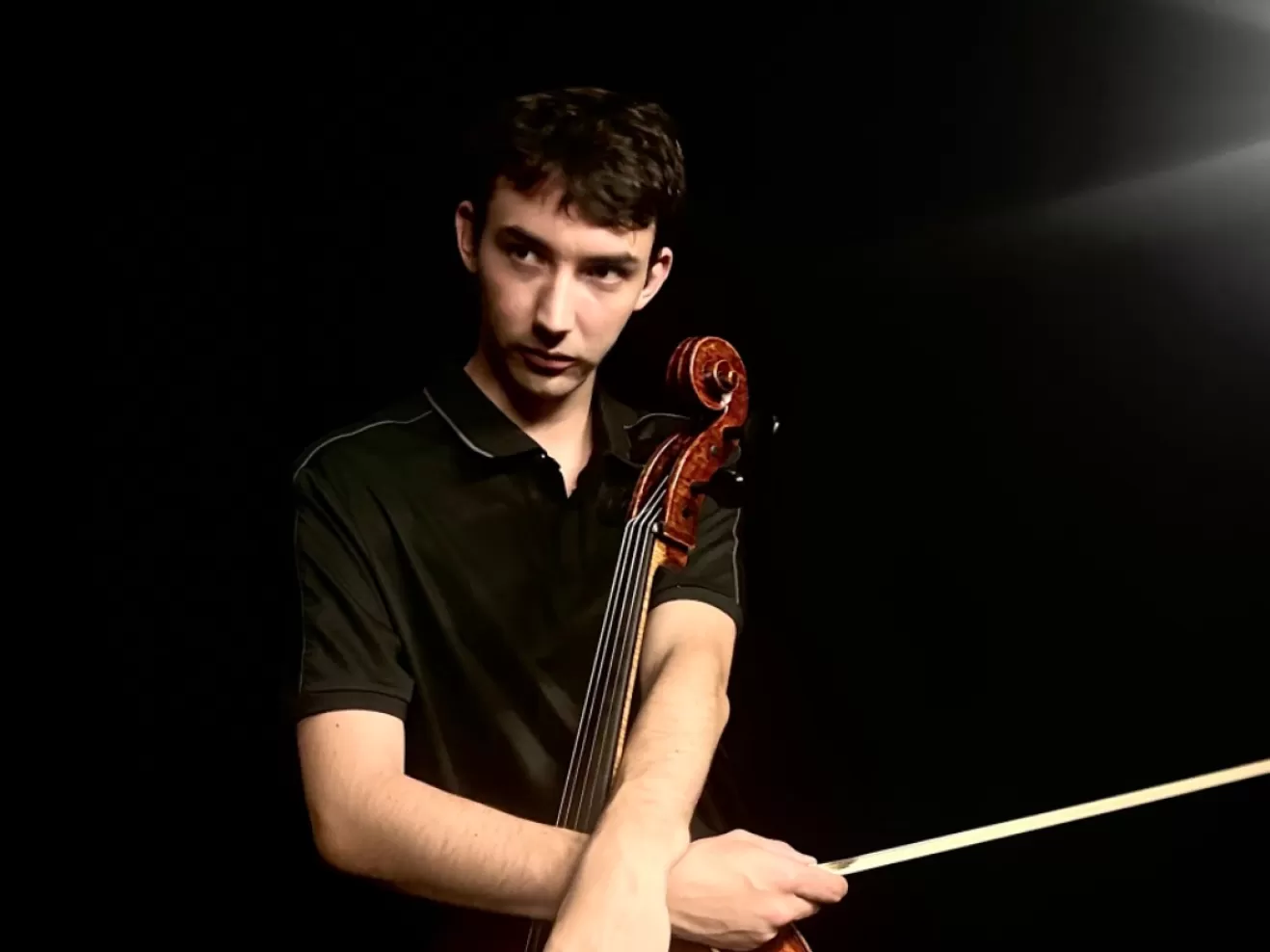 Junger Mann mit Cello vor schwarzem Hintergrund, er schaut ernst zur Seite.