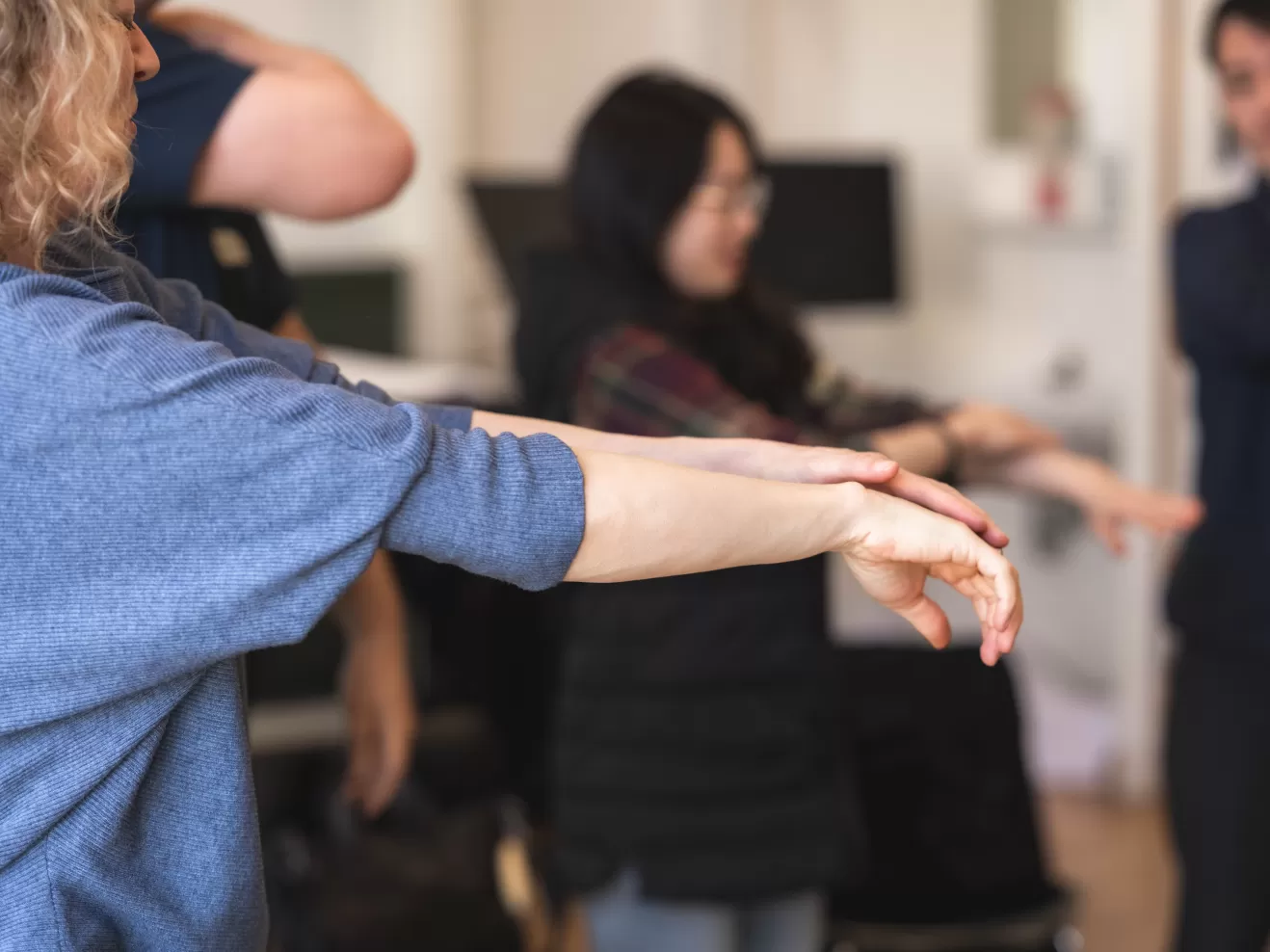 Eine Frau hat beide Arme nach vorne ausgestreckt und streicht sich mit der linken über die rechte Hand. Im Hintergrund erkennt man, dass weitere Personen im Raum es ihr gleich tun.