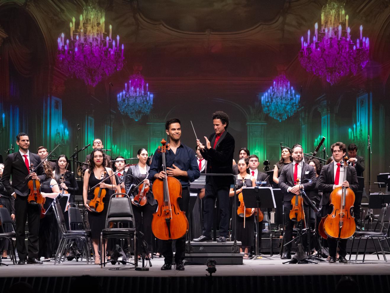 Ein Sinfonieorchester steht zum Applaus, ganz vorne in der Mitte steht lächelnd ein Cellist. Schräg hinter ihm applaudiert der Dirigent.