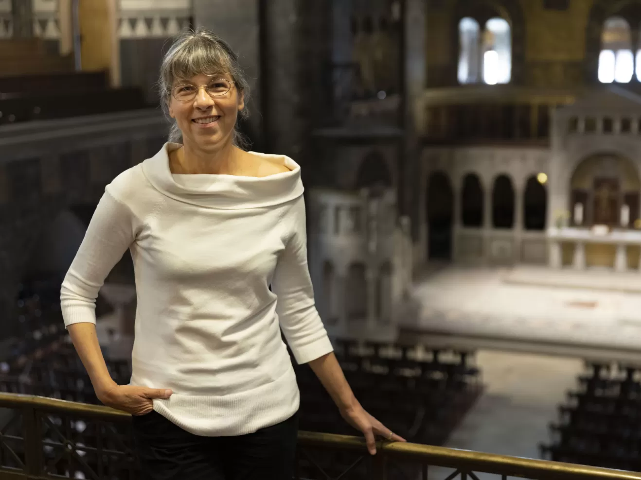 Susanne Rohn trägt ein schickes, weißes Oberteil und hält sich an der Emporenbrüstung fest. Blick in Kirchenraum