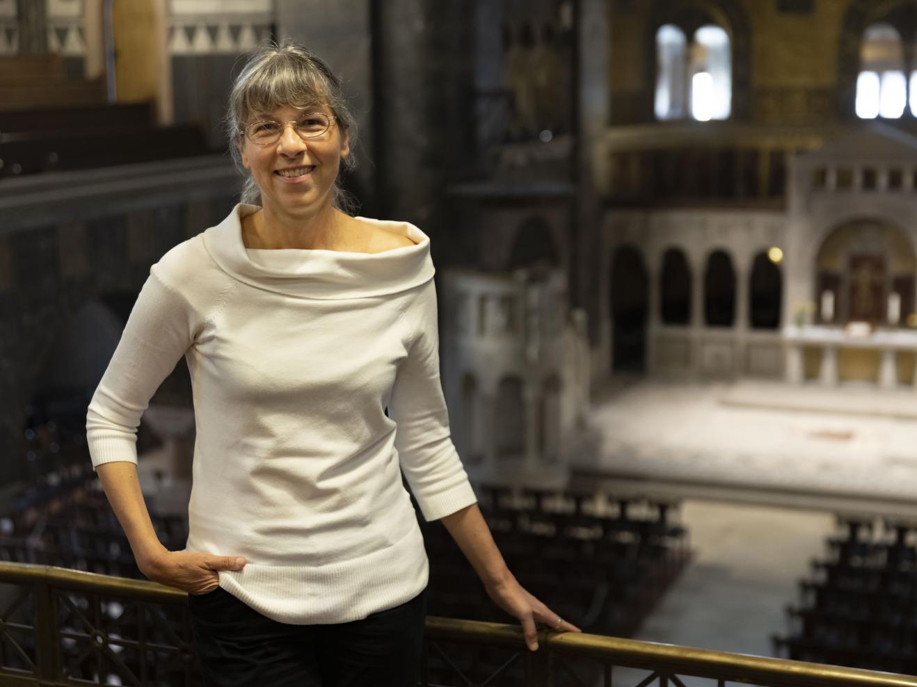 Susanne Rohn trägt ein schickes, weißes Oberteil und hält sich an der Emporenbrüstung fest. Blick in Kirchenraum