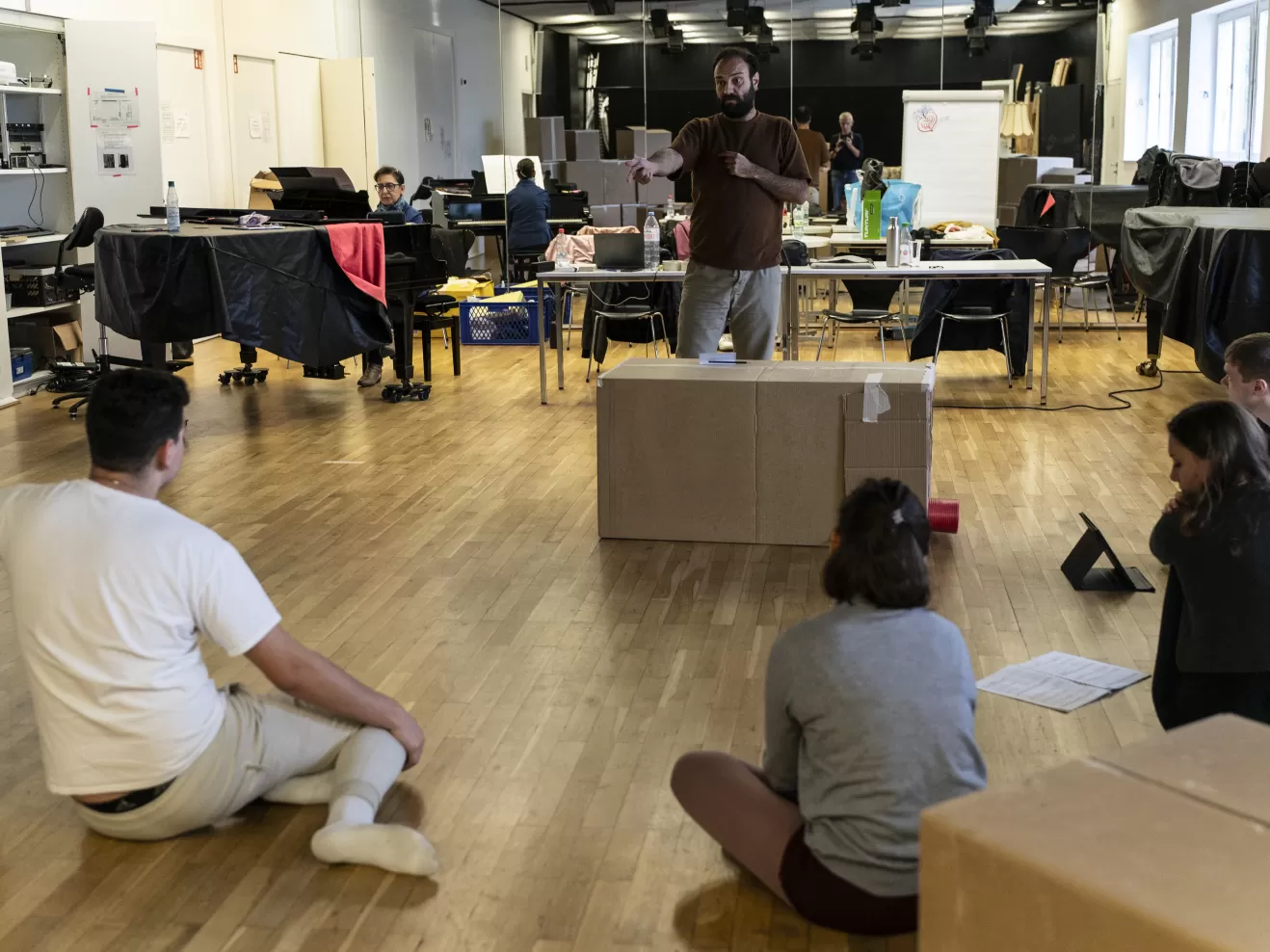 Proben im Opernstudio, ein Mann gibt Regieanweisungen, während die Darstellenden auf dem Boden sitzen
