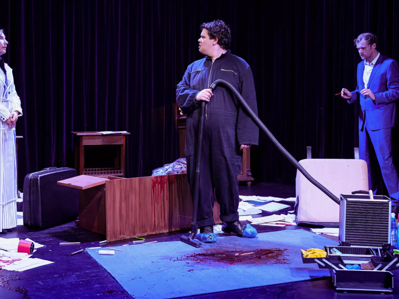 Drei Personen stehen in einem Raum. Die Person in der Mitte hält einen Staubsauger und schaut zur linken.