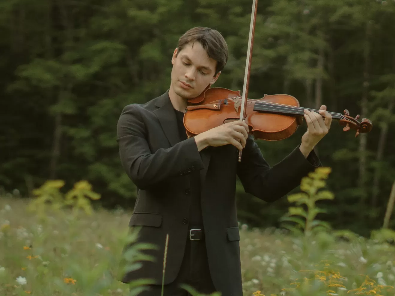 Ein junger Mann in einem schwarzen Anzug steht in einer Wiese und spielt mit geschlossenen Augen auf einer Bratsche