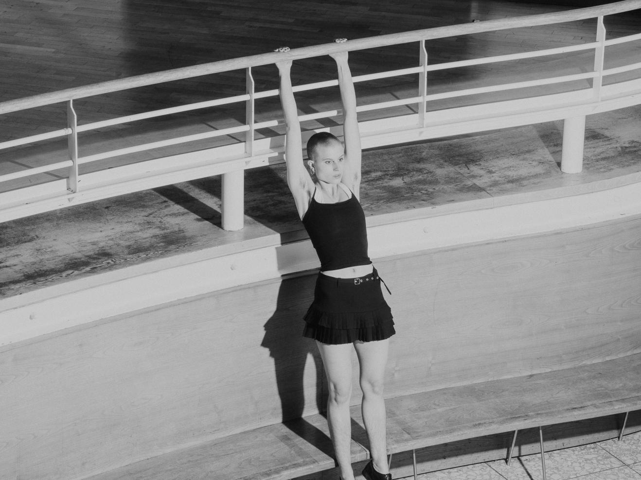 Magazin Schwarz-weiß Bild einer jungen Frau, die im Foyer der HfMDK an einem Geländer hängt, wie an einer Turnstange.