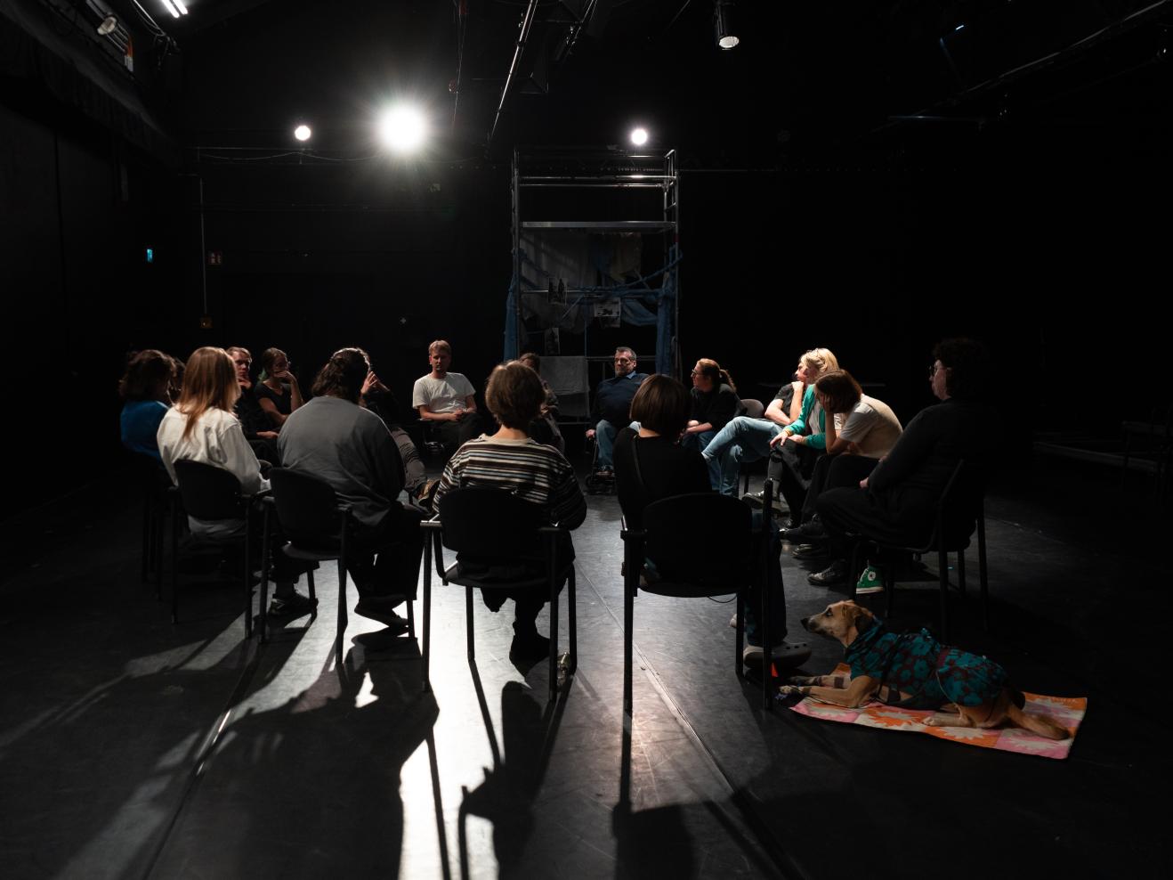 Personen sitzen in einem Stuhlkreis auf einer Bühnenfläche.