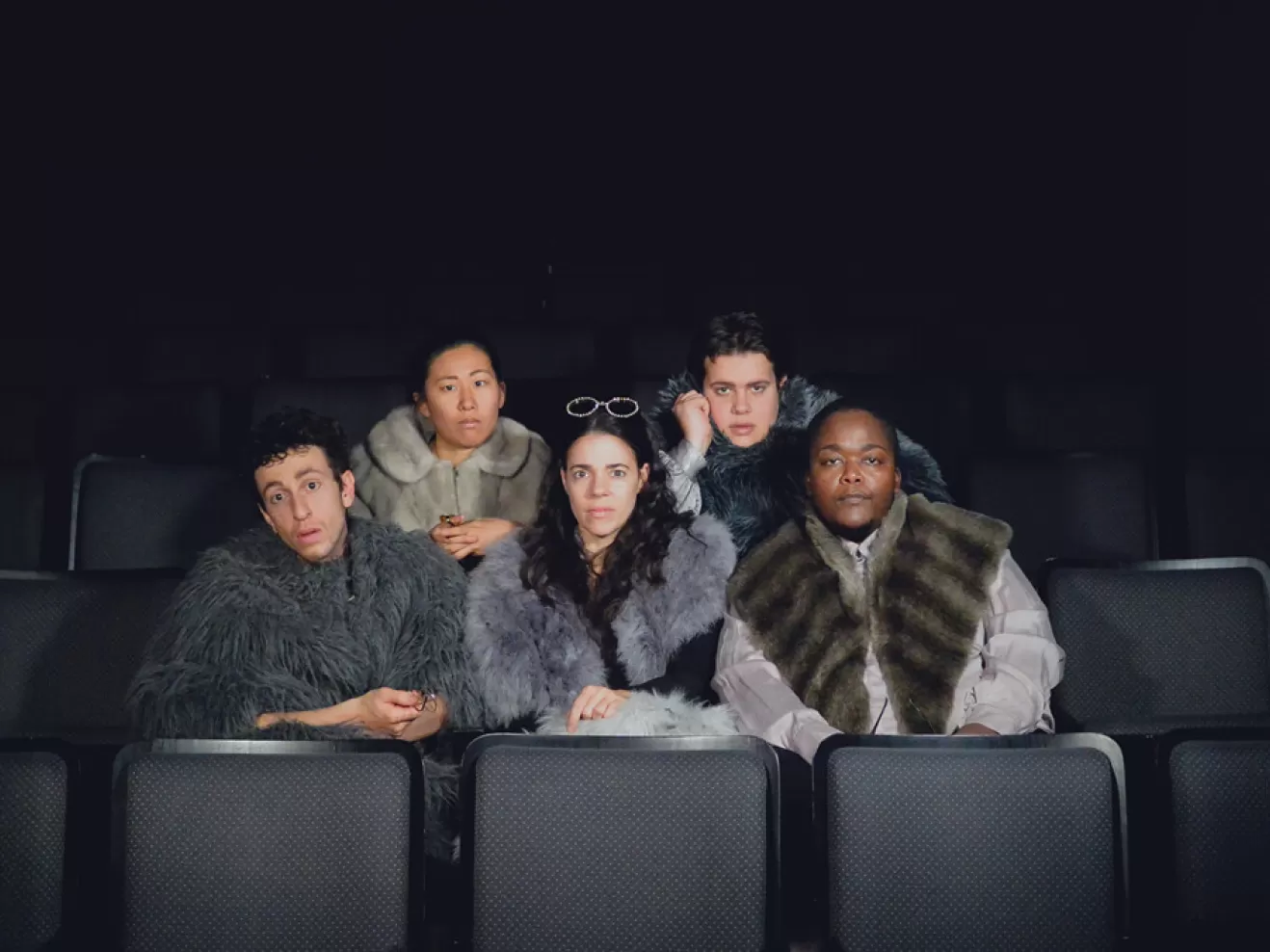 Mehrere Leute sitzen auf schwarzen Sitzen und tragen graue Klamotten