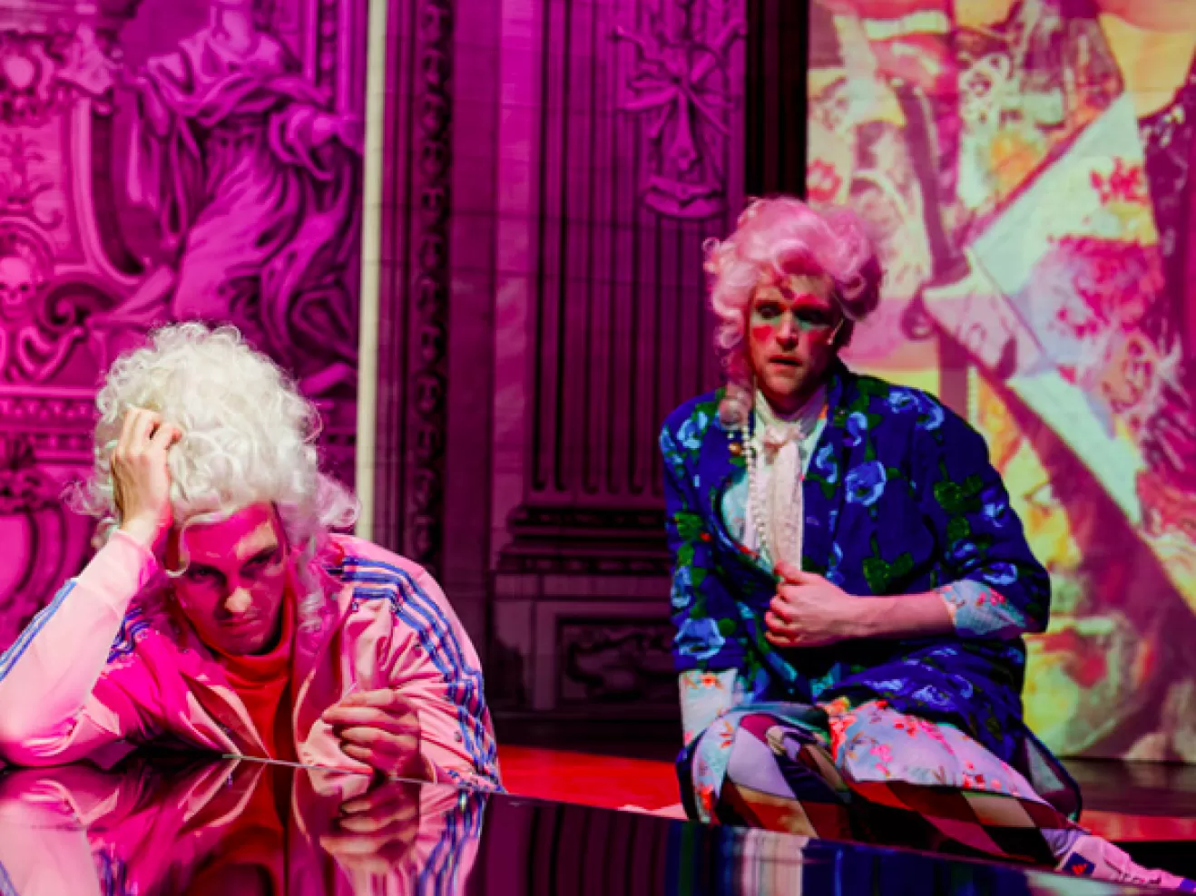 Zwei Menschen sind bunt angezogen und sitzen auf einer pinken Bühne.