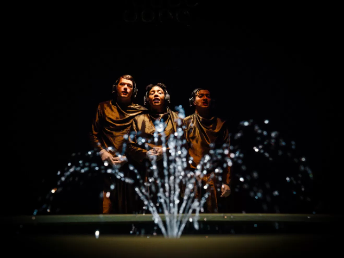 Drei Menschen tragen goldene Anzüge und stehen hinter einer Fontäne