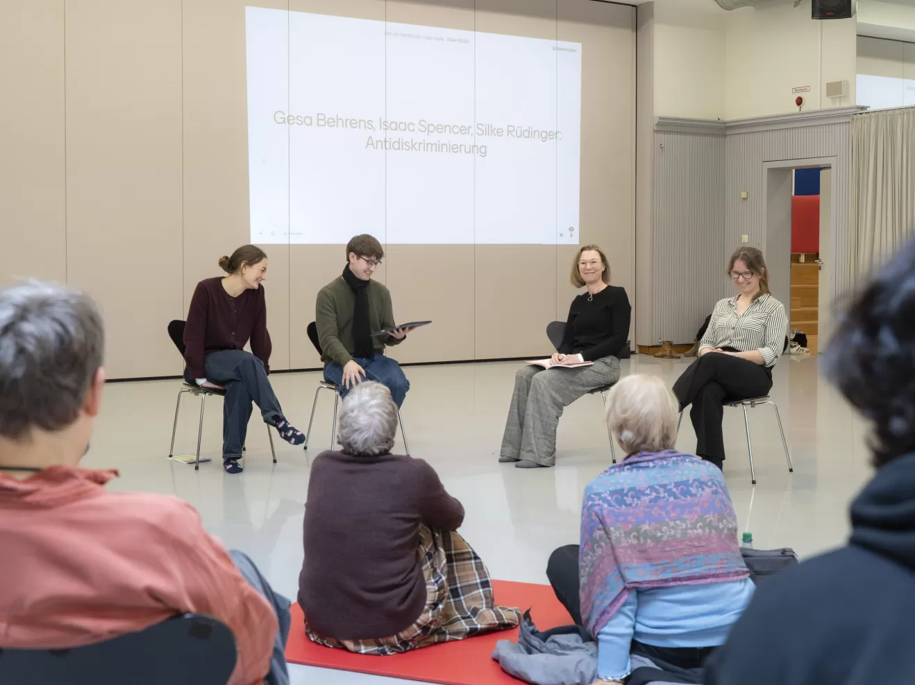 Vier Personen sitzen vor einer Gruppe Zuhörer*innen 