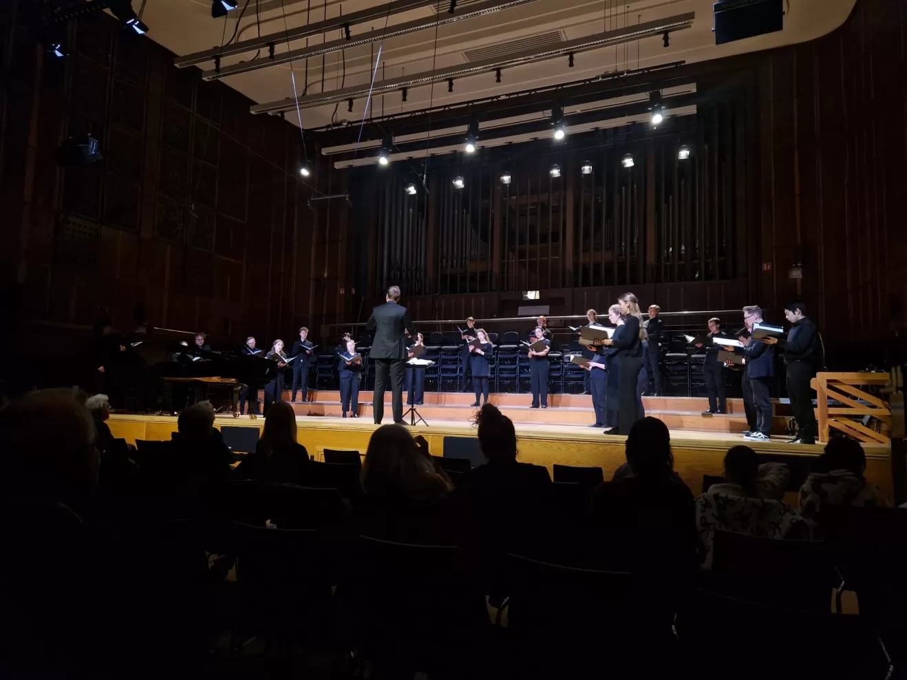 Blick in Großen Saal, der Chor steht mit etwas Abstand zueinander auf der Bühne und singt. Ein Student steht mittig und dirigiert.